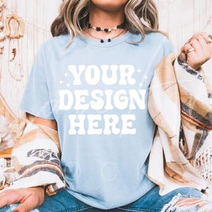 May include: A light blue t-shirt with the text "YOUR DESIGN HERE" in white. The shirt is being worn by a person with long blonde hair and a brown and white plaid blanket draped over their shoulders.