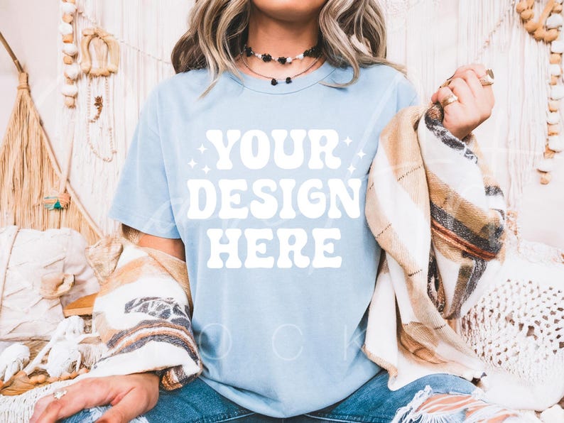May include: A light blue t-shirt with the text "YOUR DESIGN HERE" in white. The shirt is being worn by a person with long blonde hair and a brown and white plaid blanket draped over their shoulders.