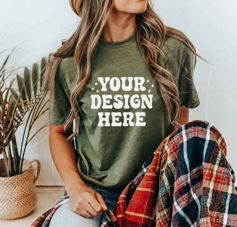 以下が含まれることがあります： 白い文字で「YOUR DESIGN HERE」と書かれた緑色のTシャツを着た女性。彼女は、チェック柄の毛布を膝にかけて座っている。