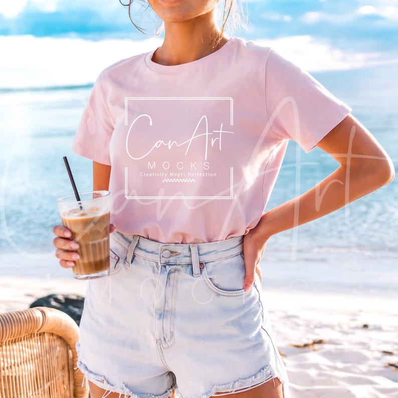 May include: A woman wearing a pink t-shirt with the text "Can Art Mocks Creativity Meets Perfection" on the front. She is standing on a beach and holding a glass of iced coffee.