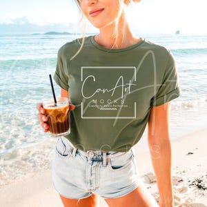 May include: A woman wearing a green t-shirt with the text "CanArt MOCKS Creativity Meets Perfection" on the front. She is standing on a beach and holding a glass of iced coffee.
