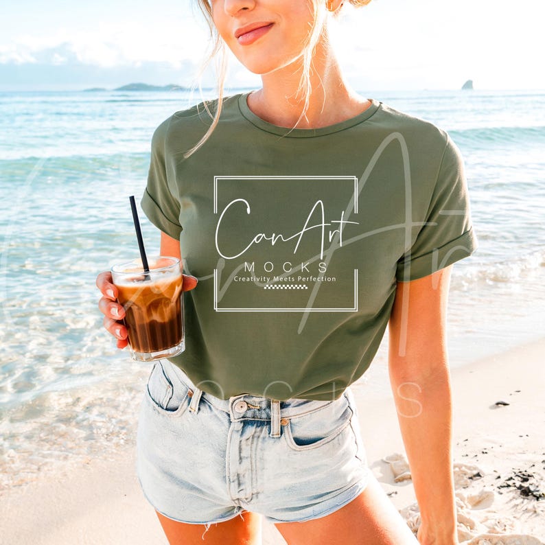 May include: A woman wearing a green t-shirt with the text "CanArt MOCKS Creativity Meets Perfection" on the front. She is standing on a beach and holding a glass of iced coffee.