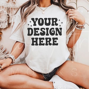 May include: A white t-shirt with the text "YOUR DESIGN HERE" in black retro-style lettering. The shirt is being worn by a person sitting on a white surface.