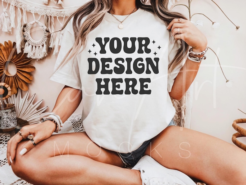 May include: A white t-shirt with the text "YOUR DESIGN HERE" in black retro-style lettering. The shirt is being worn by a person sitting on a white surface.