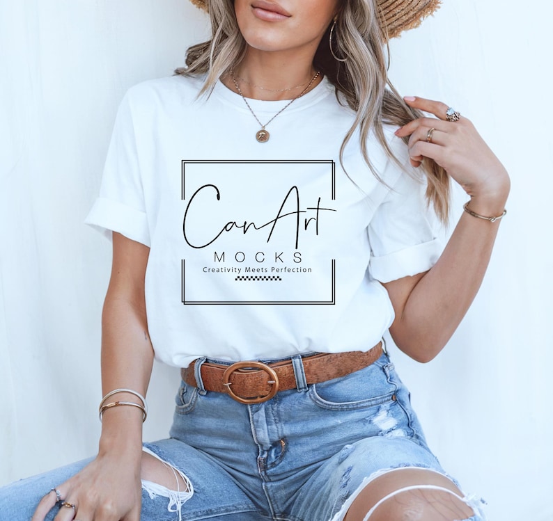 Pode incluir: Uma camiseta branca com um gr&aacute;fico preto que diz "CanArt" e "MOCKS Creativity Meets Perfection". A camiseta est&aacute; sendo usada por uma pessoa com cabelo longo e loiro e um chap&eacute;u de palha. A pessoa est&aacute; usando jeans azul com um cinto marrom.