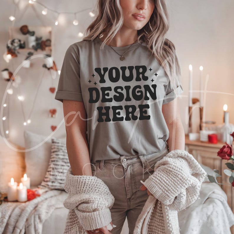 May include: A gray t-shirt with the text "YOUR DESIGN HERE" in black. The shirt is being worn by a person in a cozy setting with string lights and candles.