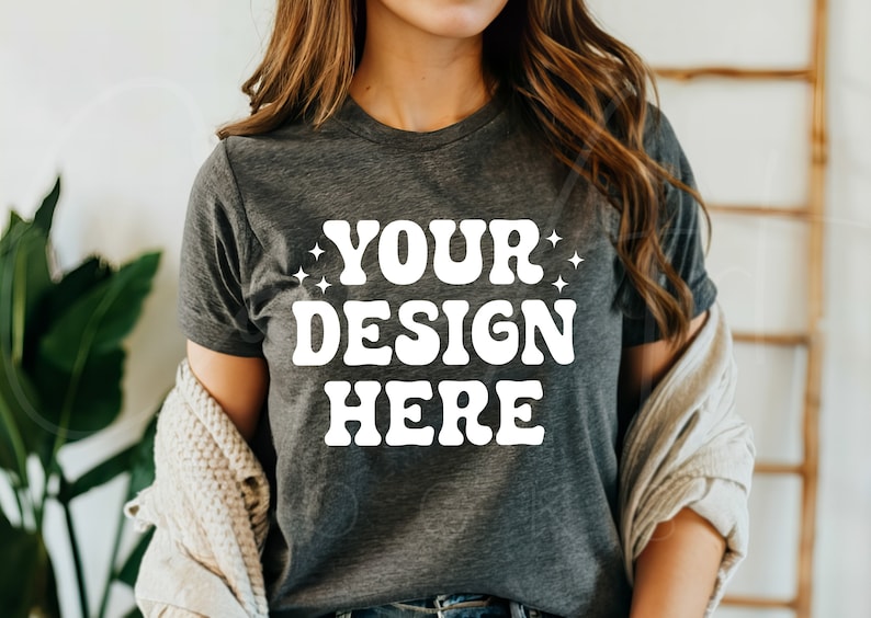 Op de afbeelding: Een donkergrijs T-shirt met de tekst "YOUR DESIGN HERE" in wit, retro-stijl lettertype. Het shirt wordt gedragen door een persoon met lang bruin haar.