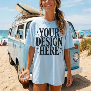 Könnte beinhalten: Hellblaues T-Shirt mit dem Text "YOUR DESIGN HERE" in Schwarz, mit Sternakzenten. Im Hintergrund ein blauer Vintage-Van mit Surfbrett. Die Person hält einen transparenten Becher mit einem braunen Getränk.