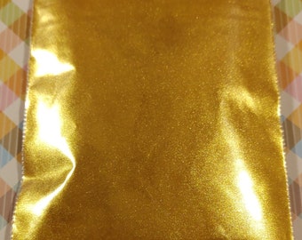 May include: A sealed, clear plastic bag filled with fine, shimmering gold glitter. The glitter reflects light, creating a sparkling effect. The bag is on a patterned surface.