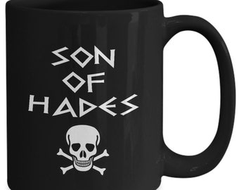 May include: Black ceramic mug with the words "SON OF HADES" in white, stylized lettering. Below the text is a white skull and crossbones graphic. The mug has a curved handle.