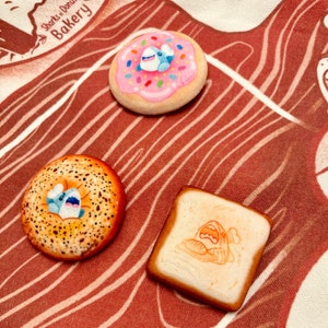 May include: Three hand-painted food-themed magnets: a pink donut with sprinkles and a blue shark, a bagel with a shark, and a slice of bread with a bread-themed graphic. The magnets are on a red and white patterned fabric.