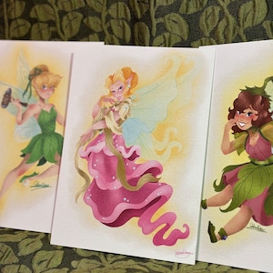 Peut inclure: Trois impressions d'art de fées à l'aquarelle. Les fées sont représentées avec des traits détaillés, portant des robes et des vêtements en forme de feuilles. La palette de couleurs comprend des verts, des roses et des jaunes. Le fond est un tissu vert texturé.