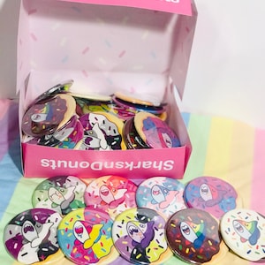 May include: A pink box with the text "SharksnDonuts" contains several round, colourful buttons. Each button features a cartoon shark design within a donut shape, decorated with sprinkles. The buttons are displayed on a rainbow striped surface.
