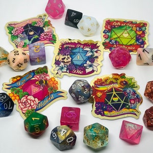 May include: A collection of colorful polyhedral dice and decorative stickers. The dice are various colors, including pink, green, and blue, with gold numbering. The stickers feature floral designs and phrases like "I'm all Natural" and "Play With Pride".