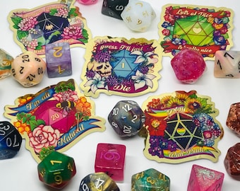 May include: A collection of colorful polyhedral dice and decorative stickers. The dice are various colors, including pink, green, and blue, with gold numbering. The stickers feature floral designs and phrases like "I'm all Natural" and "Play With Pride".