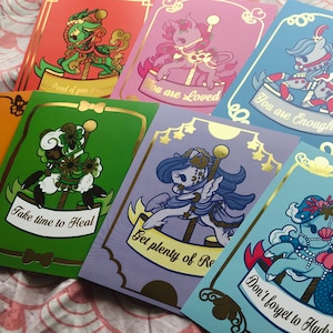 May include: A collection of colorful greeting cards with gold foil accents. Each card features a unique carousel-themed illustration with encouraging messages like "You are Loved" and "Take time to Heal". The cards are on a pink and white patterned surface.
