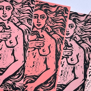 May include: A collection of pink and black linocut prints. Each print features a nude female figure with long flowing hair, her hand gently touching her chest. The prints are displayed in a fan-like arrangement.