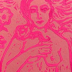 May include: A pink and peach-colored print of a woman's portrait. The artwork features a woman with long flowing hair and a hand placed on her chest. The image has a textured, carved appearance, with a floral element in the upper left corner.