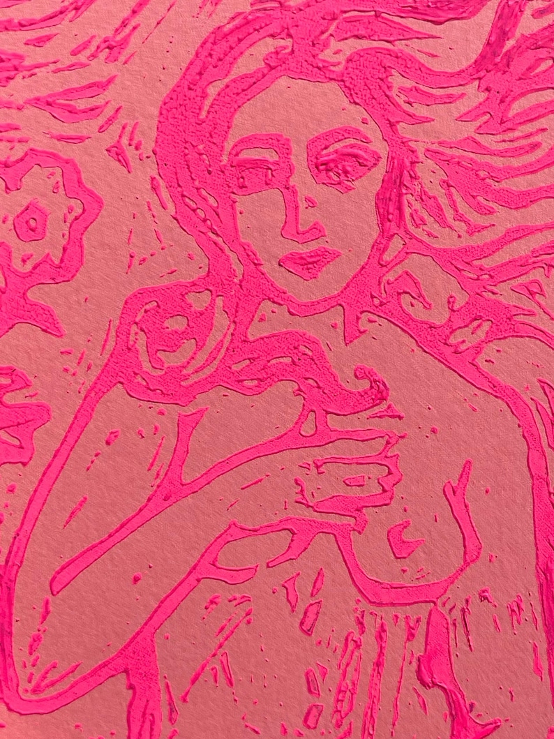 May include: A pink and peach-colored print of a woman's portrait. The artwork features a woman with long flowing hair and a hand placed on her chest. The image has a textured, carved appearance, with a floral element in the upper left corner.