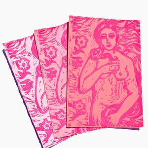 May include: Three pink linocut prints. The prints feature floral and figure designs. The figure print depicts a nude woman with long flowing hair. The prints are in varying shades of pink and are arranged in a fan shape.