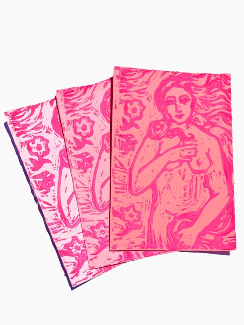 May include: Three pink linocut prints. The prints feature floral and figure designs. The figure print depicts a nude woman with long flowing hair. The prints are in varying shades of pink and are arranged in a fan shape.