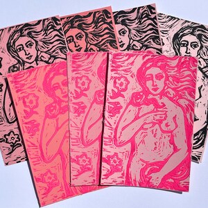 May include: A collection of pink and black linocut prints featuring a woman with flowing hair and floral elements. The prints vary in shades of pink and depict the female figure holding a flower. The art pieces are arranged on a white surface.