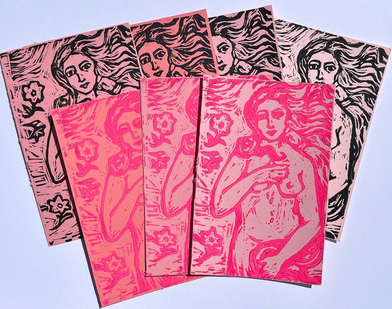 May include: A collection of pink and black linocut prints featuring a woman with flowing hair and floral elements. The prints vary in shades of pink and depict the female figure holding a flower. The art pieces are arranged on a white surface.