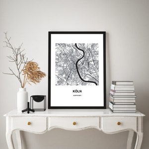 May include: Black and white city map print of Cologne, Germany, with the city name and coordinates printed below the map. The print is framed in a black frame and is displayed on a white dresser with a vase and decorative objects.