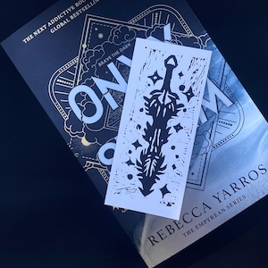 May include: A book titled "Brave the Dark" by Rebecca Yarros, part of the Empyrean series. The book cover features a gold and black design with a sword and stars. A bookmark with a black and white illustration of a sword is on top of the book.