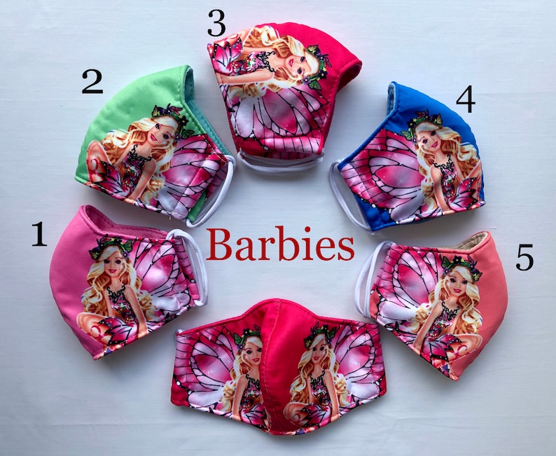 May include: Five pink, blue, and green fabric face masks with a white elastic band. Each mask features a different Barbie fairy design with pink wings and glitter. The masks are numbered 1 through 5.