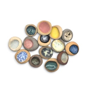 May include: A collection of fifteen small, round, hand-formed ceramic stones with concave centers. Each piece features a unique glazed interior in various colors including pink, yellow, blue, green, and speckled patterns, set against a natural tan clay exterior.