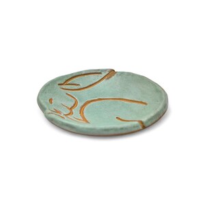 May include: A small, round, ceramic dish in a light teal color. The dish features a carved design of a sleeping rabbit in a warm brown color. The rabbit is lying down with its ears folded back. The dish is a decorative item.