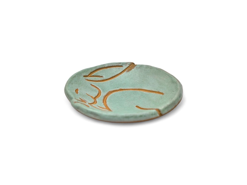 May include: A small, round, ceramic dish in a light teal color. The dish features a carved design of a sleeping rabbit in a warm brown color. The rabbit is lying down with its ears folded back. The dish is a decorative item.