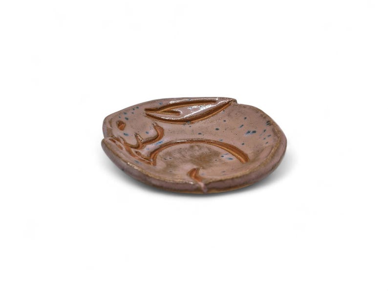 May include: A handmade ceramic dish in the shape of a rabbit. The dish is a mottled brown color with blue speckles and a raised rabbit design. The dish is ideal for holding small items.