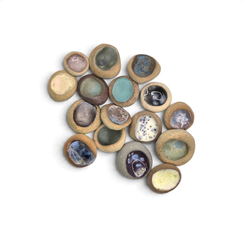 May include: A collection of fifteen small, rounded ceramic stones with a textured, tan-colored exterior and smooth, concave centers featuring various glossy glazes in shades of blue, purple, white, green, and brown, arranged in a cluster.