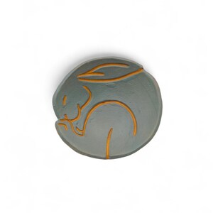 May include: A round, gray ceramic dish with an orange outline of a sleeping rabbit. The rabbit is curled up in a ball, with its ears and body clearly defined by the orange lines. The dish is a decorative item.