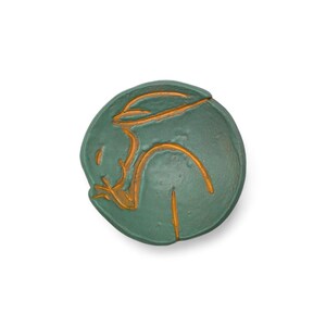 May include: A round, teal-colored ceramic dish with a gold-colored abstract design. The design appears to be a stylized human figure. The dish is set against a white background.