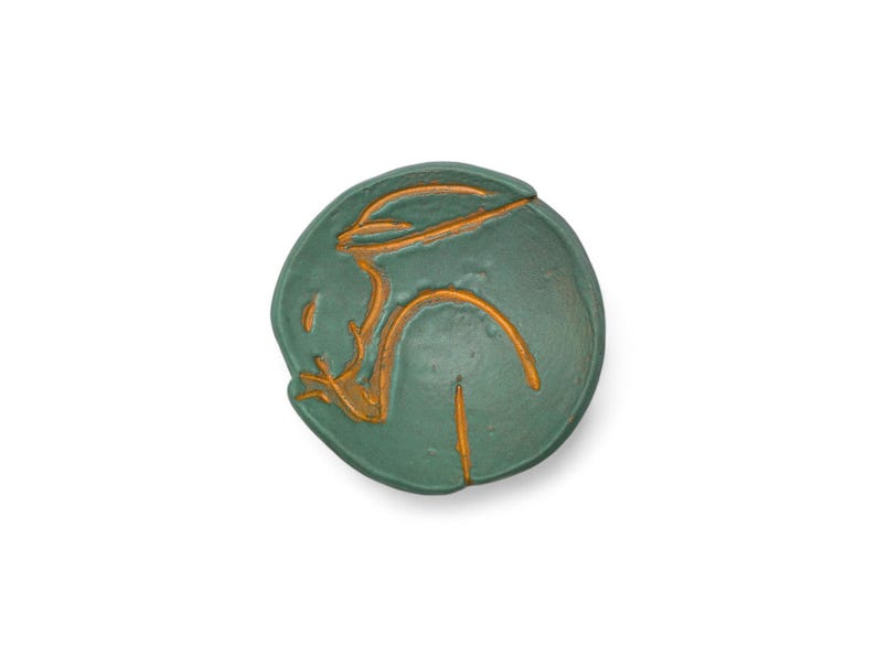 May include: A round, teal-colored ceramic dish with a gold-colored abstract design. The design appears to be a stylized human figure. The dish is set against a white background.