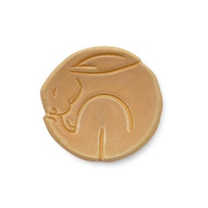 May include: A tan-colored, circular ceramic piece featuring a sleeping rabbit design. The rabbit is curled up, with its ears and body forming a continuous line. The design is embossed on the surface, creating a subtle relief.