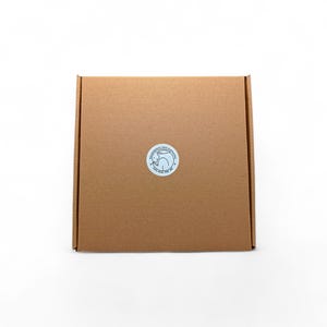 May include: A square, brown cardboard box with a circular sticker on the front. The sticker features a blue and white design with text. The box is centered on a white background.
