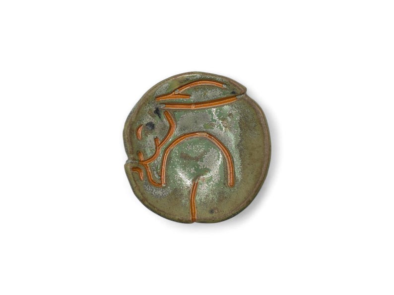 May include: A ceramic dish with a green glaze and a rabbit design in orange. The rabbit is depicted in a stylized, minimalist form. The dish has a rounded shape and is set against a white background.