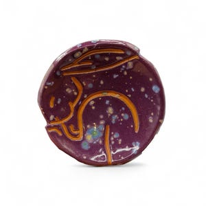 May include: A round, ceramic decorative piece with a deep purple glaze and a speckled pattern. The design features an orange outline of a rabbit. This unique art piece is perfect for home decor.