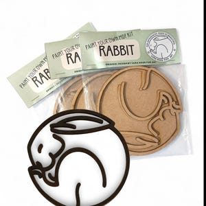 May include: A craft kit featuring a wooden rabbit design. The kit includes several laser-cut wooden rabbits and packaging labeled "Paint Your Own MDF Kit Rabbit." The design includes a white rabbit with a brown outline.