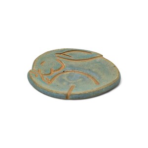 May include: A ceramic, round, blue-green coaster with a carved rabbit design. The rabbit is depicted in a sleeping pose, with its ears and body outlined in a lighter brown color. The coaster is a handmade item.