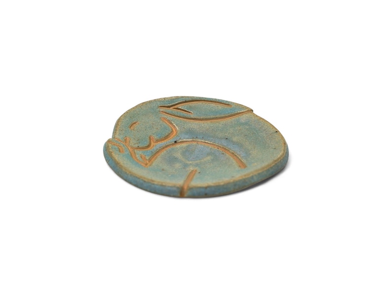 May include: A ceramic, round, blue-green coaster with a carved rabbit design. The rabbit is depicted in a sleeping pose, with its ears and body outlined in a lighter brown color. The coaster is a handmade item.