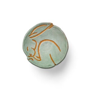 May include: A round, ceramic dish in a soft green hue, featuring a sleeping rabbit design in a contrasting golden-brown color. The rabbit is curled up, with its ears and body outlined. The dish is likely handmade.