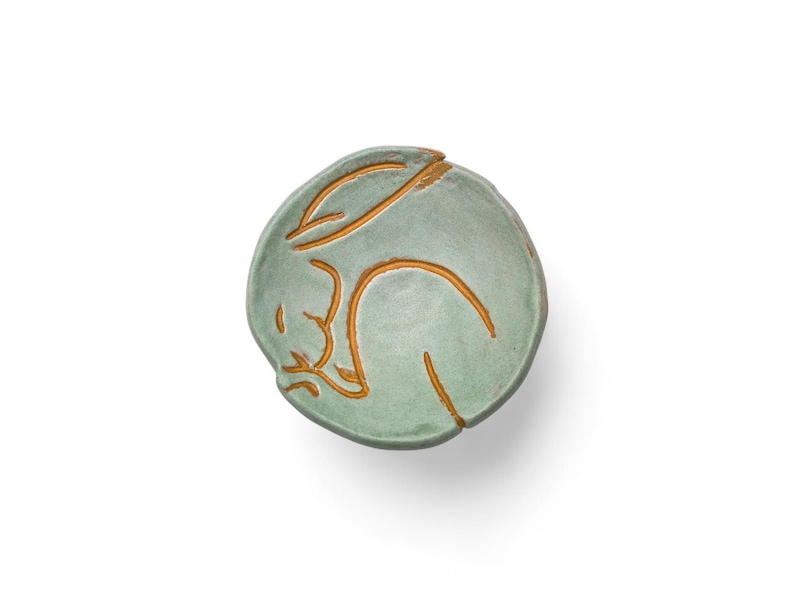 May include: A round, ceramic dish in a soft green hue, featuring a sleeping rabbit design in a contrasting golden-brown color. The rabbit is curled up, with its ears and body outlined. The dish is likely handmade.