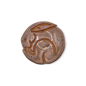 May include: A ceramic bunny figurine in a circular shape. The bunny is curled up, with its ears and body outlined in a darker brown. The base color is a light brown with blue speckles.