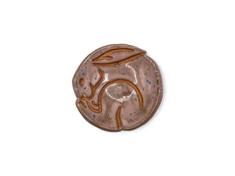 May include: A ceramic bunny figurine in a circular shape. The bunny is curled up, with its ears and body outlined in a darker brown. The base color is a light brown with blue speckles.