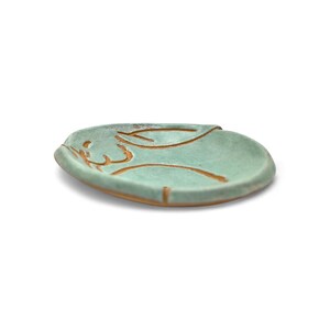 May include: A small, round, ceramic dish with a light teal glaze. The dish features a brown, abstract design on the surface. The rim of the dish has a slightly raised edge. The dish is likely handmade and suitable for holding small items.